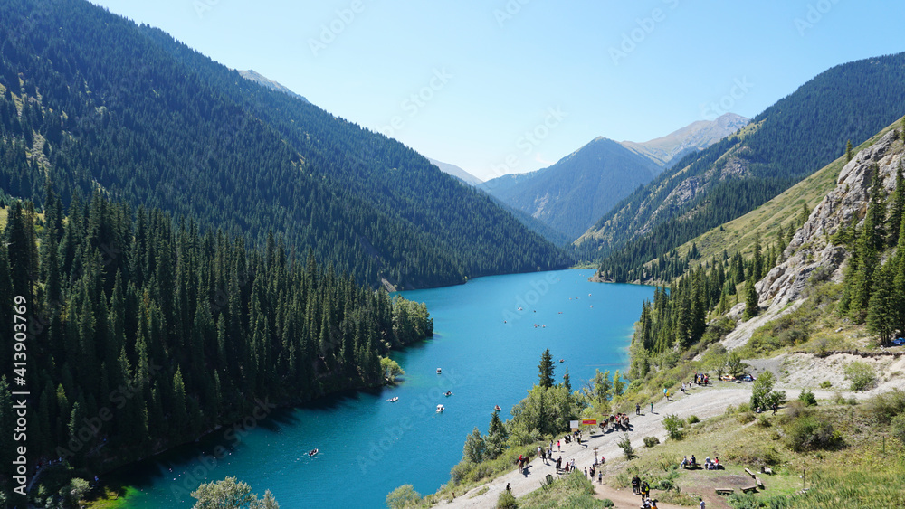 Kolsay lake among green hills and mountains. The mountain lake is surrounded by green forest, tall coniferous trees, grass and bushes. Clean water is like a mirror. Tourists swim on boats. Kazakhstan Kolsay lake among green hills and mountains. The mountain lake is surrounded by green forest, tall coniferous trees, grass and bushes. Clean water is like a mirror. Tourists swim on boats. Kazakhstan