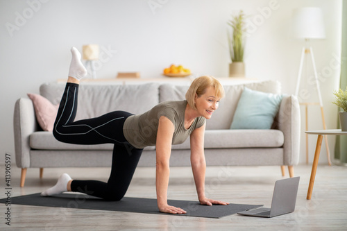 Online home fitness concept. Beautiful mature woman exercising to sports video on laptop in living room