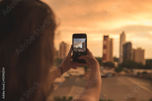 Rear view of woman photographing buildings with smart phone against sky during sunset photo