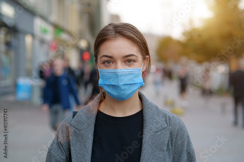 Young woman in face mask stands at crowded street and looks at the camera