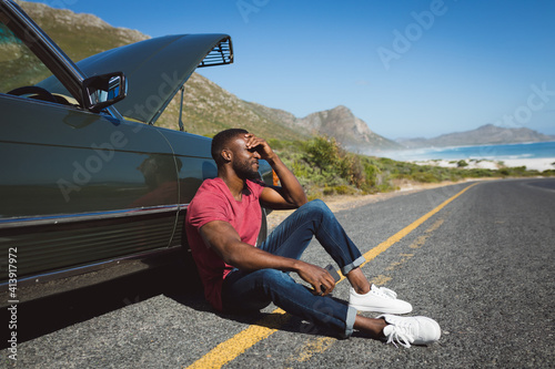 African american man holding smartphone sitting on road beside broken-down car with open bonnet