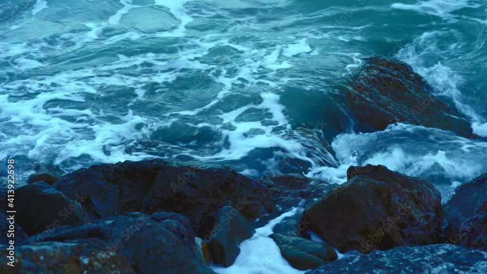 Sea water waves splashing with foam at rocky stones coast, blue color toned.