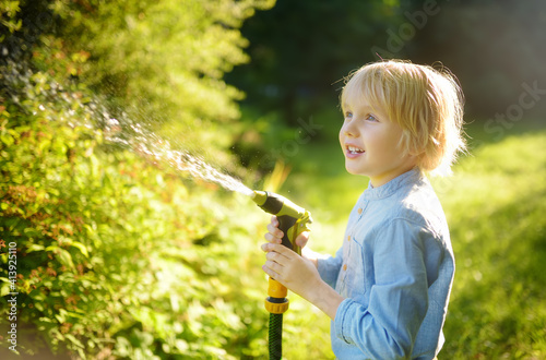 Funny little boy watering plants and playing with garden hose with sprinkler in sunny backyard. Preschooler child having fun with spray of water.