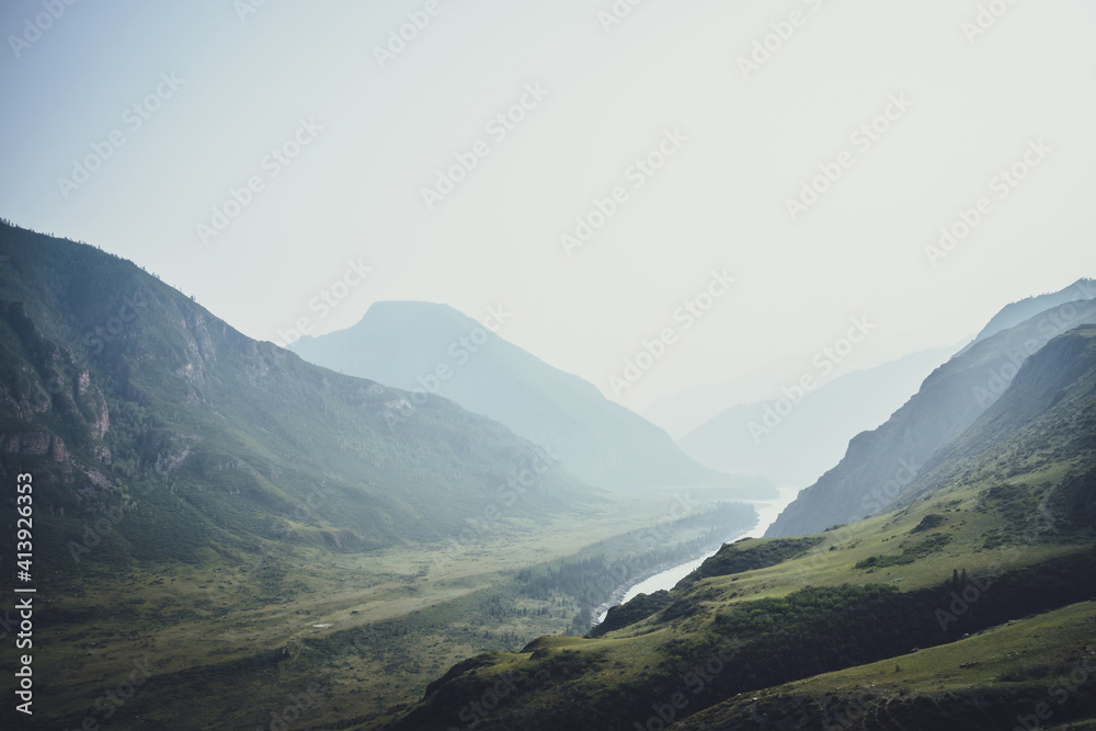 Misty mountain landscape with hills and rocks on background of wide mountain river in mist. Atmospheric scenery with mountain relief and big river in dark green valley in rainy weather. Gloomy weather