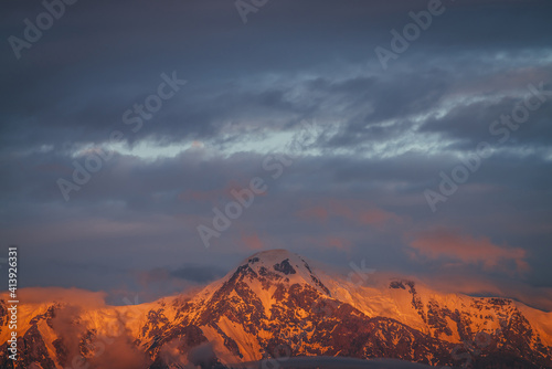 Scenic mountain landscape with great snowy mountains lit by dawn sun among low clouds. Awesome alpine scenery with high mountain pinnacle at sunset or at sunrise. Big glacier on top in orange light.