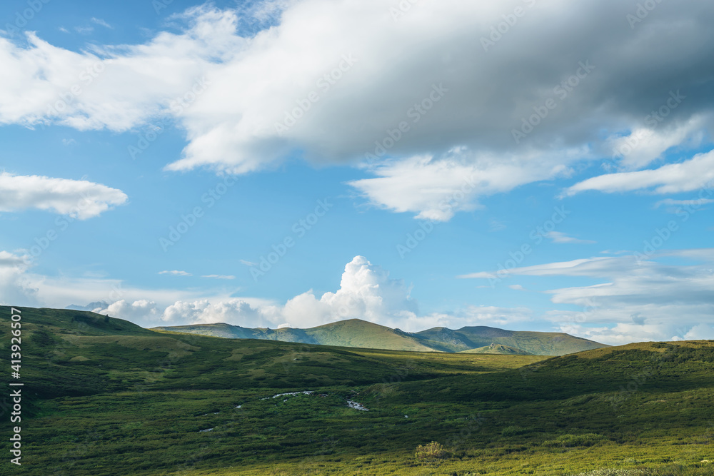 Green sunny mountain landscape with big white cloud in form of explosion in blue sky above green hills in sunlight. Beautiful sunny scenery with big cloud in shape of explosion above green mountains.