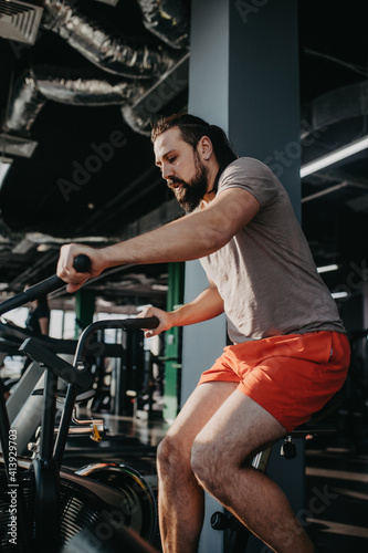 An active man with beard training in gym, riding a stationary exercise bike. Sport and quarantine.