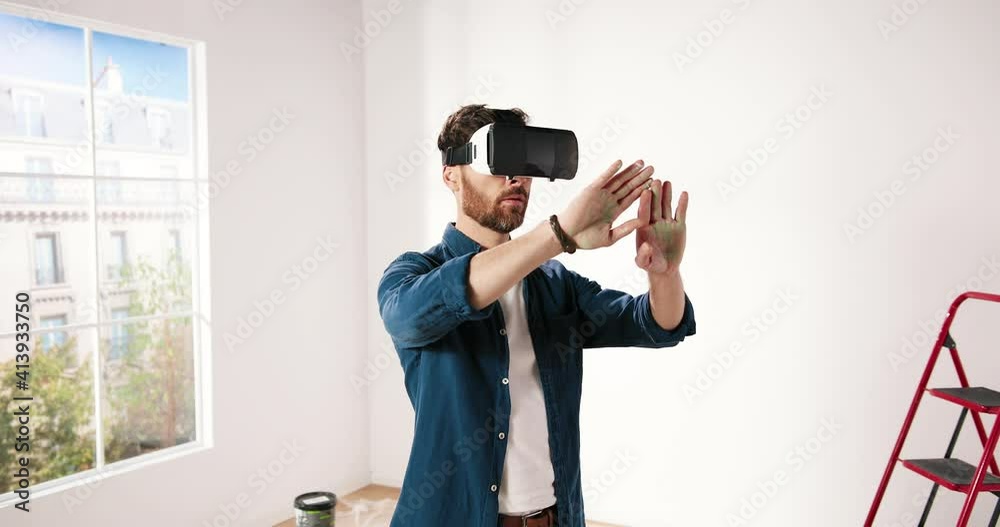 Young Caucasian bearded man in virtual reality helmet standing in new apartment and looking around using gestures.