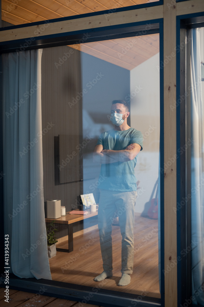 Young contemporary man in casualwear and protective mask standing by window