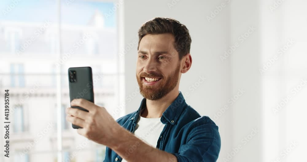 Portrait of happy caucasian man standing in new apartment on window background holding smartphone posing for selfie.