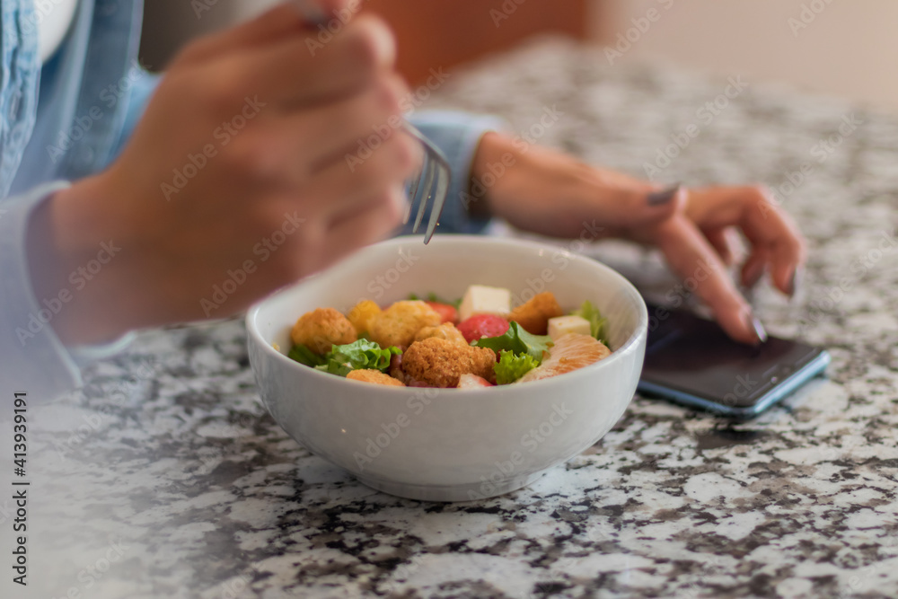 Close up of a young brunette woman eating a salad in her kitchen. She is using her phone