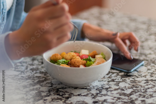 Close up of a young brunette woman eating a salad in her kitchen. She is using her phone