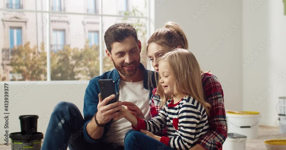 Little cute girl sits in her mother's arms and helps parents choose interior option for new room by browsing catalog on smartphone. Little cute girl sits in her mother's arms and helps parents choose interior option for new room by browsing catalog on smartphone.
