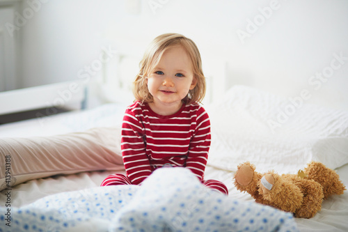 Foto Happy toddler girl in striped red and white pajamas sitting on bed right after a