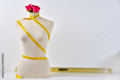 Beige sewing mannequin, on white brick background, white floor, with yellow measuring tape, colour chart, designer dress hanging on wall