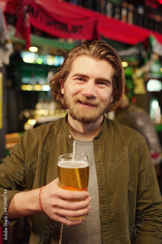 Young cheerful bearded man looking at you with smile while toasting with beer