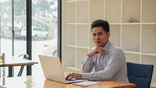 Image of a young man sitting at a desk in an office with his hands, feet, chin and smiles.