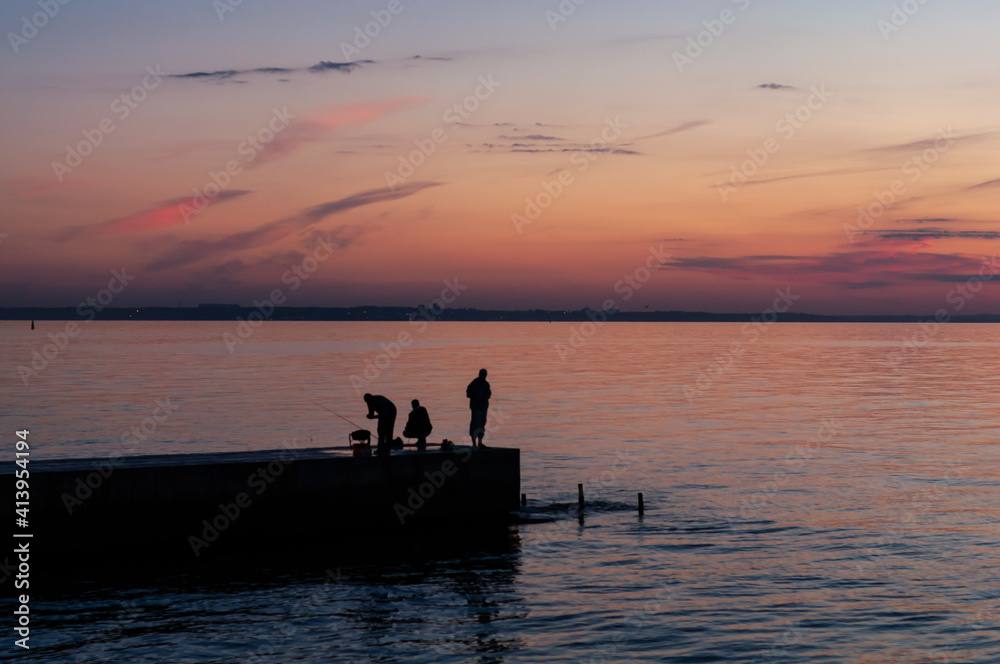 Fishing on sea, colorful morning sunrise, people on pier fish, red-orange sunrise, smooth water surface.