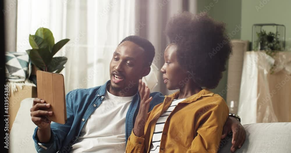 Young beautiful african american couple in new apartment browsing online catalog on tablet sitting on sofa on background of packed boxes. Young beautiful african american couple in new apartment browsing online catalog on tablet sitting on sofa on background of packed boxes.