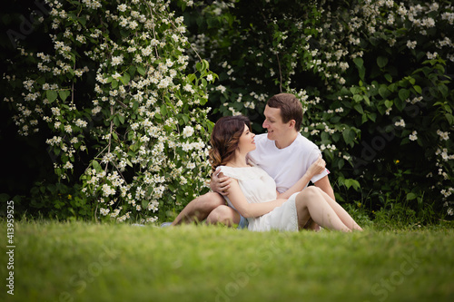 Young man and woman couple in a blooming garden near jasmine bush. Tender holding each other. Spring love story. Brown-haired girl with long hairs with her boyfriend or husband. Young couple on a date
