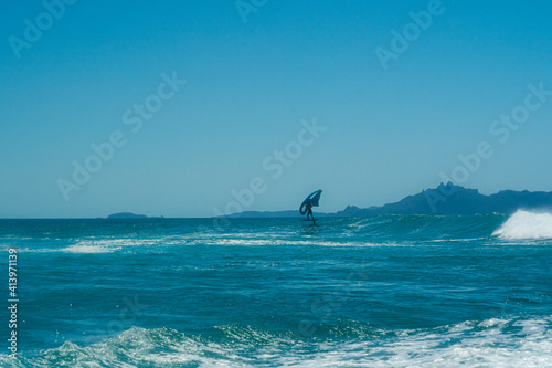 A man wing foils standing on a hydrofoil surfboard, using a hand held wing in a beautiful turquoise sea with clear blue sky and an island in the distance.