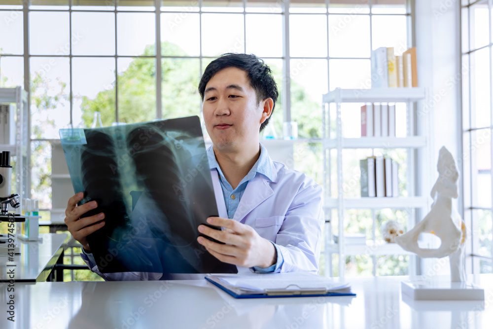 Asian male doctor sitting in hospital cabin reading and analysing issue in lungs of patient with x-ray film while wearing a labcoat Asian male doctor sitting in hospital cabin reading and analysing issue in lungs of patient with x-ray film while wearing a labcoat
