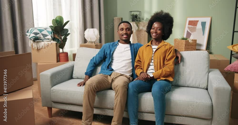 Joyful young family couple sitting on sofa smiling after unpacking and decorating new apartment with many boxes. African American couple wife and husband resting after relocation. settle in concept Joyful young family couple sitting on sofa smiling after unpacking and decorating new apartment with many boxes. African American couple wife and husband resting after relocation. settle in concept