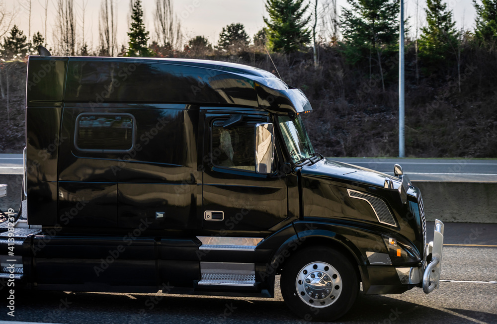 Fototapeta premium Black industrial shiny big rig semi truck with grille guard and sleeping compartment in the cab transporting cargo driving on the highway road