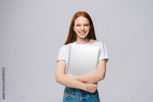 Cheerful young woman student holding laptop computer and looking at camera on isolated gray background. Pretty lady model with red hair emotionally showing facial expressions in studio, copy space.