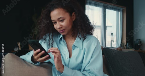 Happy confident young beautiful African American student woman using smart phone shopping online app at home on sofa. photo