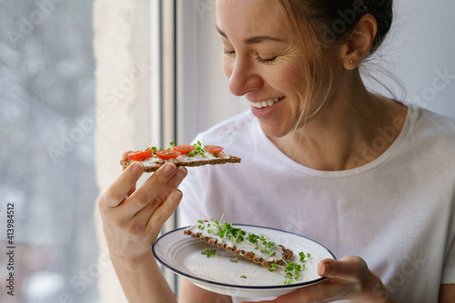 Tela Smiling woman eating rye crisp bread with creamy vegetarian cheese tofu, cherry tomato and rucola micro greens, sitting at home and looking at window
