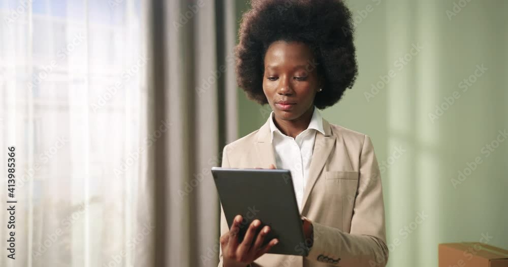 Camera approaching happy beautiful African American female real estate agent standing in empty apartment and typing on tablet device. Realtor concept. Selling property business. Close up Camera approaching happy beautiful African American female real estate agent standing in empty apartment and typing on tablet device. Realtor concept. Selling property business. Close up