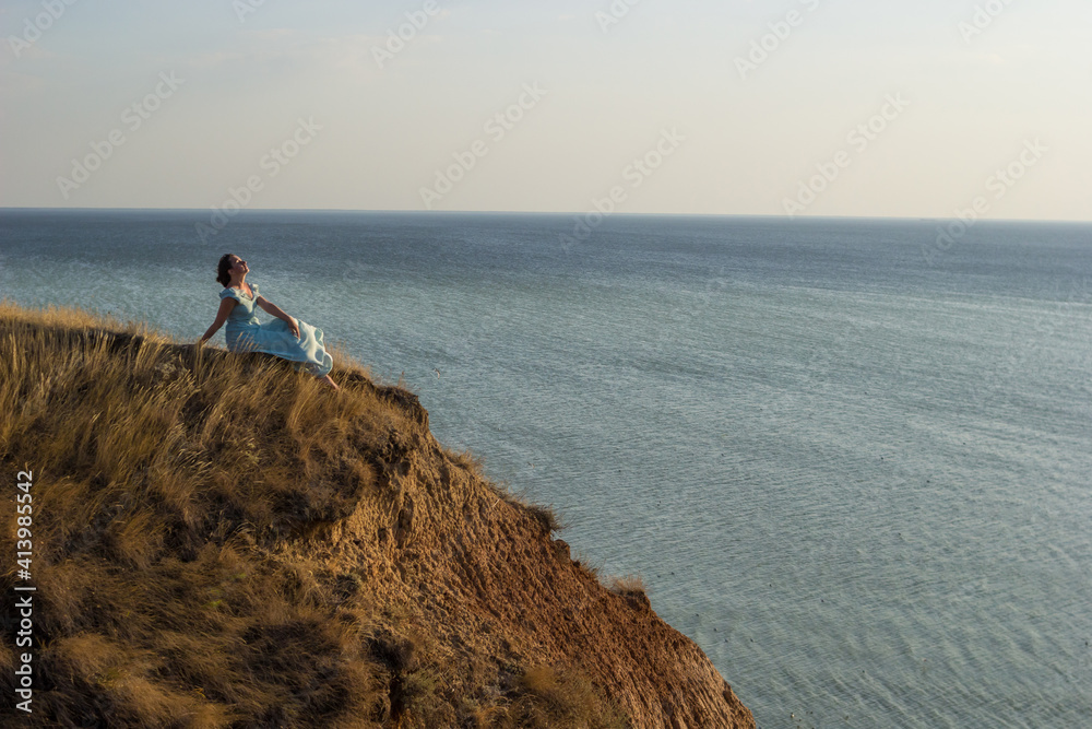 A pretty girl in a long blue dress sits on a high cliff by the sea or ocean on sunny windy summer day. Young beautiful woman with short hair watching sunset on the shores of the calm sea.