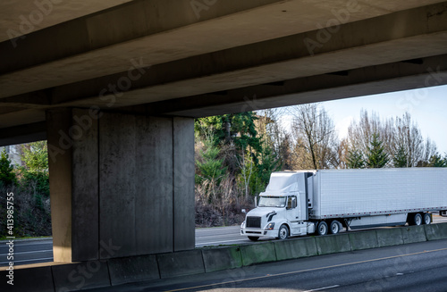 Big rig white car hauler semi truck transporting cars on the two level hydraulic modular semi trailer running on the wide divided highway with transportation bridge and overpass road intersection