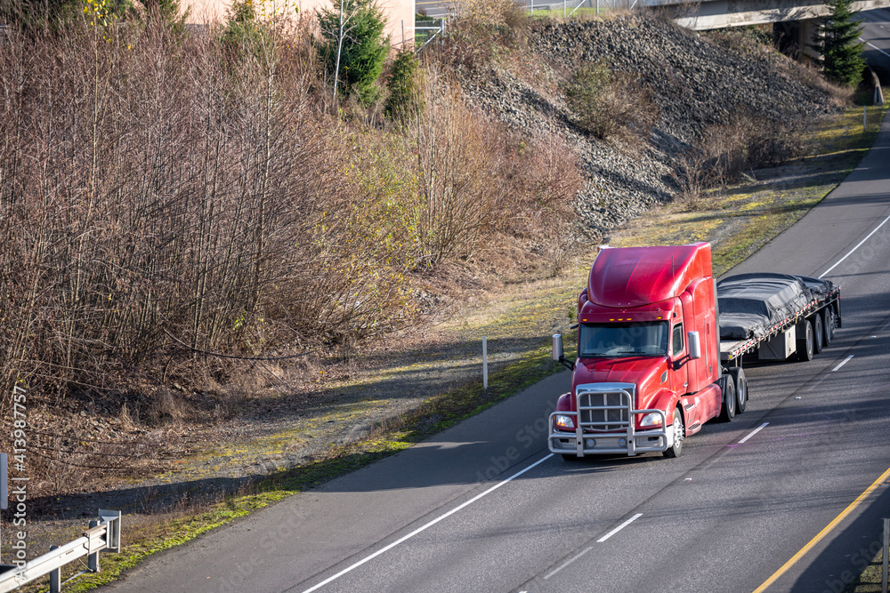 Powerful big rig red semi truck with grille guard transporting covered cargo on flat bed semi trailer driving on the turning highway road with bare winter trees on the hill