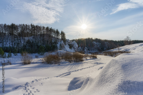 A frozen river valley with rocks and forest on its banks on a frosty winter morning. The white winter sun is shining on a blue sky with white clouds. A light fog fell on the ground due to severe frost