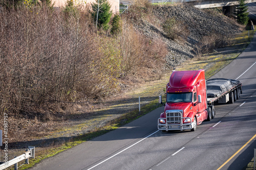Powerful big rig red semi truck with grille guard transporting covered cargo on flat bed semi trailer driving on the turning highway road with bare winter trees on the hill