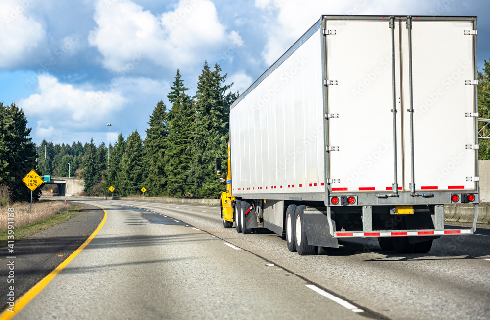 Yellow bonnet big rig semi truck transporting cargo in dry van semi trailer running on the turning wide highway road with evergreen trees on the side and bridge ahead Yellow bonnet big rig semi truck transporting cargo in dry van semi trailer running on the turning wide highway road with evergreen trees on the side and bridge ahead