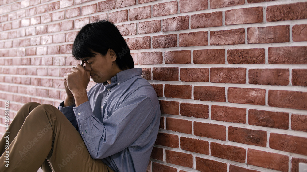 Christian Asian man is sitting against a wall, feeling sad, holding a cross and praying to God. Christian Asian man is sitting against a wall, feeling sad, holding a cross and praying to God.