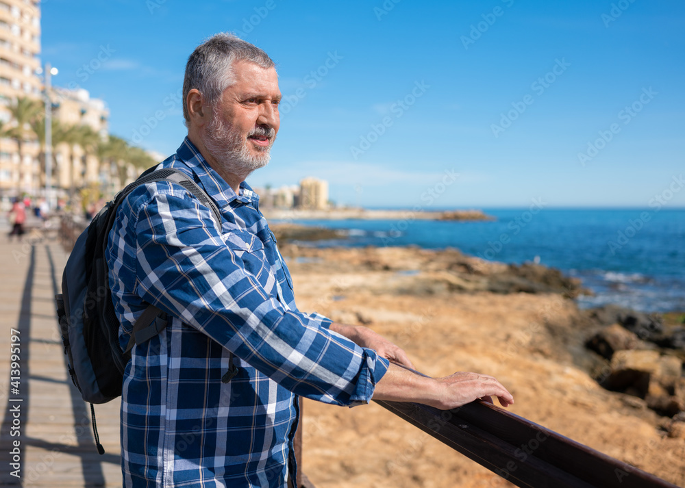 An elderly man stands in Torrevieja and looks out over the Mediterranean. He is wearing a small backpack and has his hands on the wooden railing. In the background is the promenade and the stone bank.