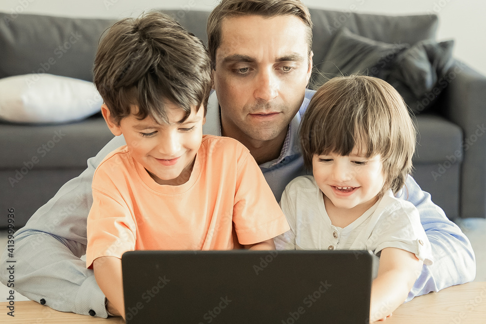 Front view of dad hugging sons and watching movie on laptop. Two lovely little boys sitting at table with father, looking at screen and smiling. Fatherhood, childhood and digital technology concept Front view of dad hugging sons and watching movie on laptop. Two lovely little boys sitting at table with father, looking at screen and smiling. Fatherhood, childhood and digital technology concept