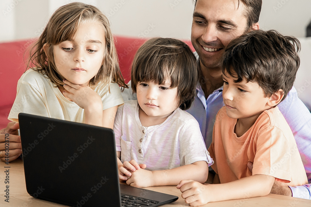 Happy dad and kids watching movie via laptop together. Caucasian father sitting at table and embracing cute children. Boys and girl looking at screen. Fatherhood and digital technology concept