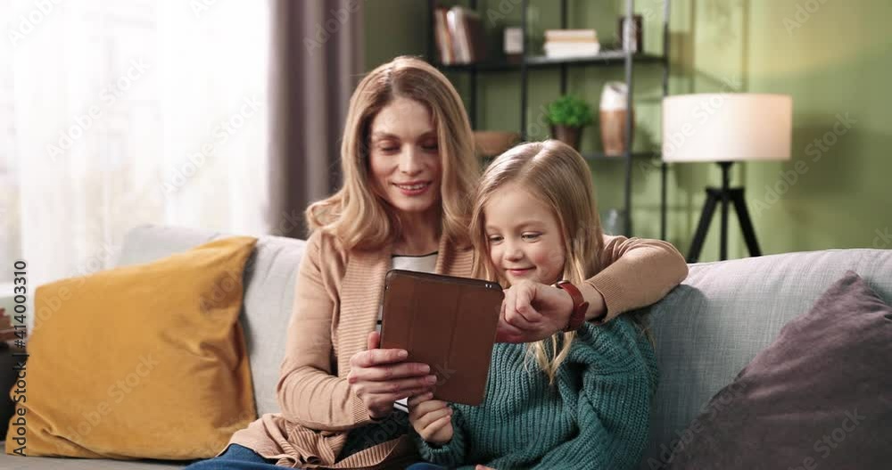 Portrait of happy smiling Caucasian beautiful mom spends time with her little cute daughter girl typing and searching internet on tablet device together sitting on sofa at home. Family concept. Portrait of happy smiling Caucasian beautiful mom spends time with her little cute daughter girl typing and searching internet on tablet device together sitting on sofa at home. Family concept.