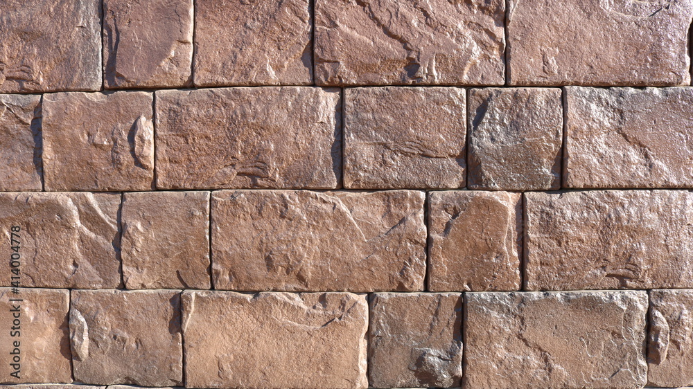 Fototapeta premium surface lined with rough natural stone of red-brown color with preserved relief texture, stone backdrop of the fragment's exterior in sunlight, emphasizing the details of cracks and bulges