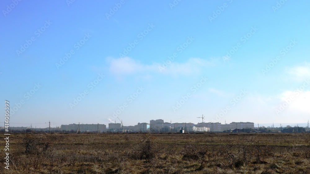 desert field with a silhouette of the city in the distance on a sunny day, a view of the outskirts of the city from the side of an empty meadow with dried grass, a panorama of the city in the haze