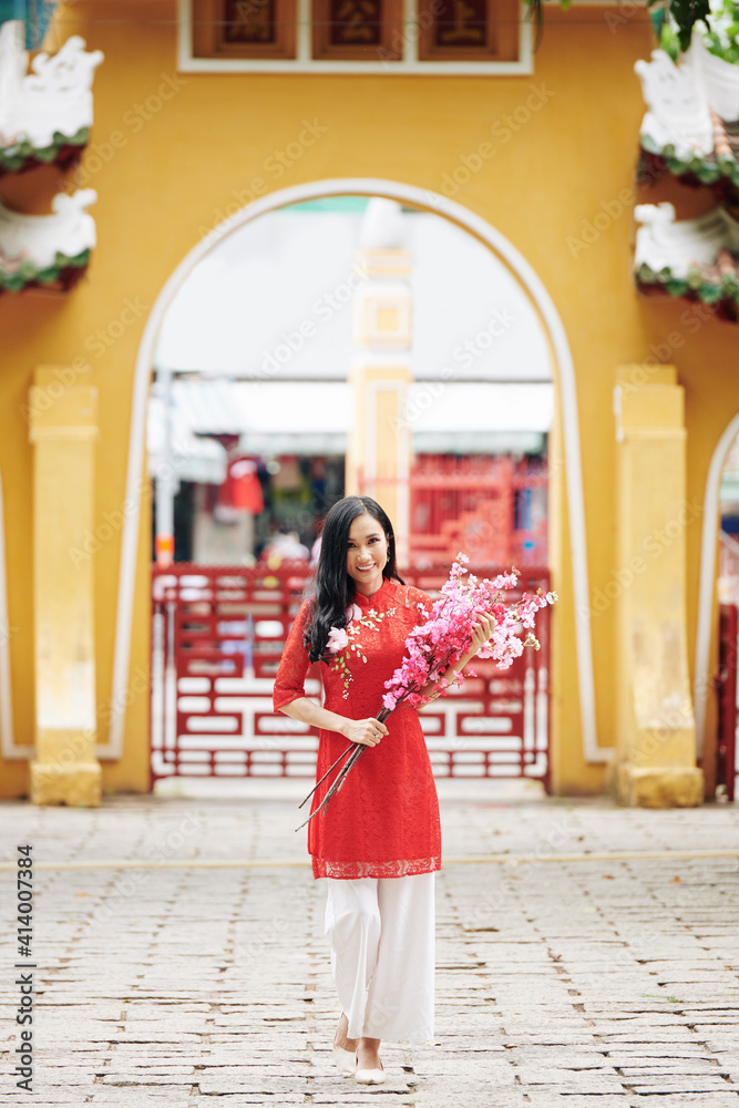 Obraz premium Attractive young Asian woman in traditional clothing walking with blooming peach branches in hands and smiling at camera