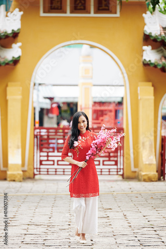 Attractive young Asian woman in traditional clothing walking with blooming peach branches in hands and smiling at camera © DragonImages