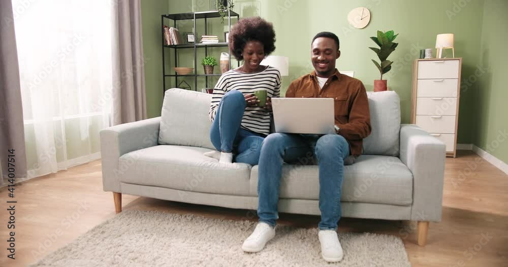 Nice lovely African American joyful married family couple man and woman sitting on sofa, resting, wife drinking hot coffee or tea and speaking in good mood while husband working typing on laptop Nice lovely African American joyful married family couple man and woman sitting on sofa, resting, wife drinking hot coffee or tea and speaking in good mood while husband working typing on laptop
