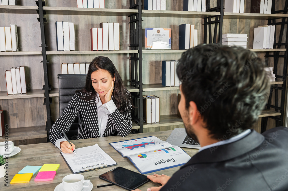 A businesswoman signing a contract document at a desk in the office sitting opposite a businessman candidate. A businesswoman signing a contract document at a desk in the office sitting opposite a businessman candidate.