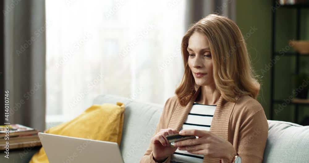 Beautiful happy blonde woman sitting at home on the couch and doing online shopping using bank card and laptop. Beautiful happy blonde woman sitting at home on the couch and doing online shopping using bank card and laptop.